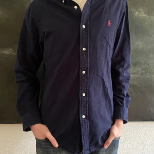 Mörkblå skjorta från Ralph Lauren - Säljer en mörkblå långärmad skjorta från Ralph Lauren med klassisk passform. Skjortan har vita knappar och röd broderad logga på bröstet. Tillverkad i mjuk bomullsflanell för extra komfort. Prsiet går såklart att diskutera!💯