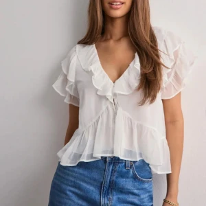 Vit volangblus med v-ringning - Säljer en luftig vit blus med v-ringning och volangdetaljer längs ärmar och hals. Blusen är tillverkad i ett tunt, lätt material och har korta ärmar. Perfekt för varma dagar och enkel att styla med jeans eller kjol.