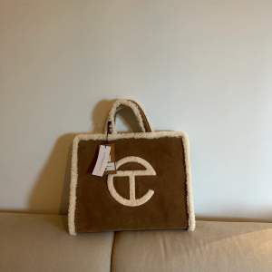 TELFAR Bag made in collaboration with UGG in genuine chestnut suede with a cuddly shearling trim. Bag features double strap to be worn cross-body, inlaid shearling TELFAR logo, 100% cotton lining with interior zipper pocket and magnetic snap closure.