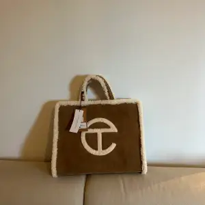 TELFAR Bag made in collaboration with UGG in genuine chestnut suede with a cuddly shearling trim. Bag features double strap to be worn cross-body, inlaid shearling TELFAR logo, 100% cotton lining with interior zipper pocket and magnetic snap closure.
