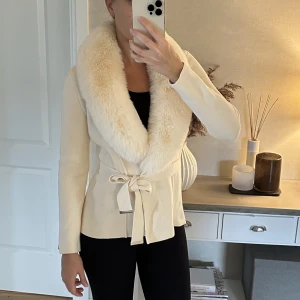 Kofta med stor pälskrage - Gräddvit, figursydd kofta med knytband och stor fluffig pälskrage från Chiara Forthi Exclusive (Bubbleroom), stl 34. Ett rejält och lyxigt plagg med snygga detaljer! Pälsen sitter fast med knappar och är därmed avtagbar. Nypris 799kr.