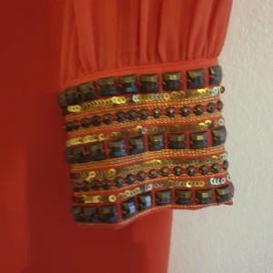 Orange fest klänning med unika detaljer - Unik klänning med.uttryckssfulla detaljer i rygg och ärmar.   Djup draperad rygg. Utsmyckad med nitar och paljetter   Köpt via köp & sälj-sida, men tyvärr fel storlek för mig. Jättefint skick. Som ny enligt min bedömnkng.