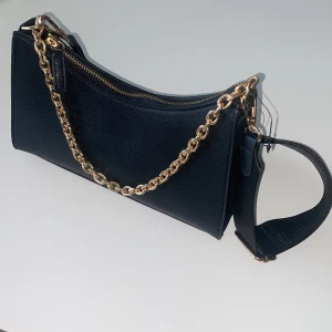 Svart axelremsväska med hängande kedja och guldiga detaljer - Väskan har aldrig använts och är helt ny. Den kan bäras på flera olika sätt som handväska eller shoulderbag. Är från Glitter.