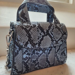 Ormskinns handväska - Handväskan är gjord at oäkta ormskinn och har en silvrig metall kedja som axelrem samt ett handtag ovanpå. Den har dessutom två olika fack på vardera sida som stängs med en magnet (se bild 2). Storlek: 18 cm bred, 12 cm lång, 8 cm djup.