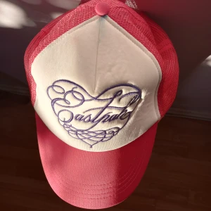 Eastpak cerise truckerkeps  - I härligt skick för den som gillar en lite inbiten keps, ceriserosa och sitter väldigt bra på! Ett snirkligt och gulligt hjärta broderat på framsidan.