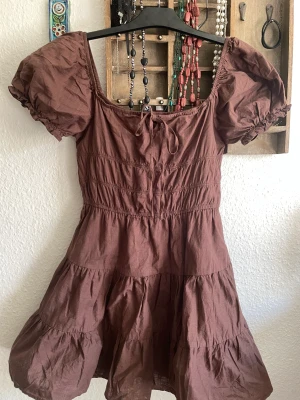 Brun klänning - Ganska kort går typ till låren ovanför knäna. Volang-illusion. resår och ”puff” i ärmarna. Resår också i midjan och knyts vid bysten