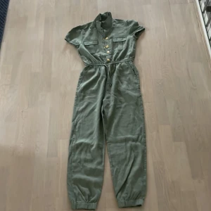 Grön byxdress - Det är en grön byxdress, den är mjuk och luftig med guldiga knappar och två fickor där fram, två fickor på sidorna, inga bakfickor. Endast använd 1 gång.