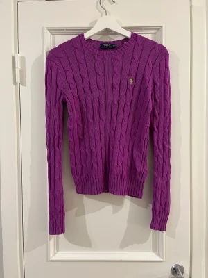 Violetfärgad kabelstickad Ralph Lauren tröja - Härlig violettfärgad Ralph Lauren POLO tröja med rund urringning. Den är i mycket bra skick och har används ett fåtal gånger. Den är i normalstorlek har en lösare passform.  Materialet är i 100% bomull och är väldigt skön på.