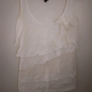 Vit ärmlös topp / linne med volanger och rosett. XS. Bomull. Lindex. - Vit ärmlös topp / linne med volanger och rosett. XS. Bomull. Lindex. Något missfärgad vid armhål men kan troligen tas bort med lite klorin.