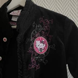 Hello Kitty tröja/kavaj/jacka i ett sammetsliknande tyg med Kitty-”emblem/broderi och knappar med rosa stenar💕  (Made in China, står storlek L men är mer som en XS) Längd 47cm. Bredd vid axlar 37 cm. Bredd under ärmarna 40 cm.  