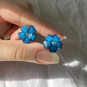 Blå blommor glittriga örhängen - Glittriga örhängen i form av blåa blommor. Bijouterier från 2000-talet, visst slitage och tecken på användning finns. Tillverkade i plast och okänd metall.