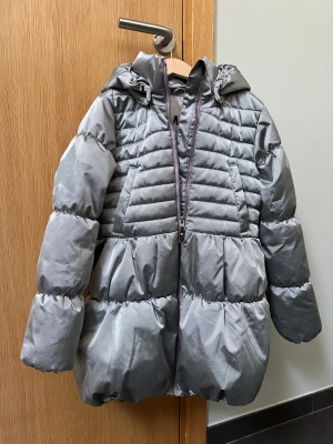 Molo vinterjacka - silvergrå - Jättefin och varm vinterjacka i storlek 140. Längre modell med fickor och avtagbar huva. Silvergrå i färgen och regnavvisande. 