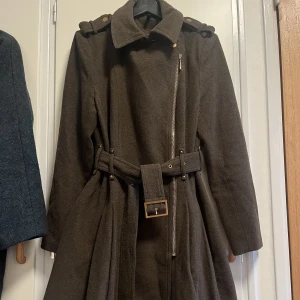 Kappa - Militärfärgad lång kappa med dragkedja, knäppning vid kragen samt skärp. Valbart hur en vill ha kragen/slaget - nedvikt eller uppknäppt. Mkt varm och 10% ull.  Sömmen har släppt lite i ryggen, se bild. Enkelt att sno ihop men hade inte mörk tråd hemma. 😆 