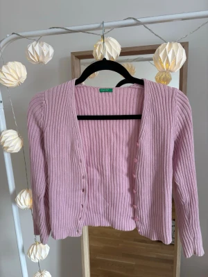 Rosa ribbad kofta med knappar - Säljer en söt rosa ribbad kofta med v-ringning och matchande knappar längs framsidan. Koftan har trekvartsarmar och är gjord i ett mjukt, stretchigt material som sitter snyggt på kroppen. Varumärke är united colors of Benetton. 