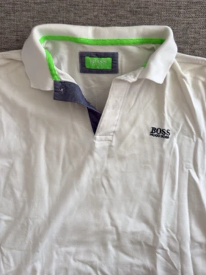 Vit pikétröja från Hugo Boss - Vit pikétröja från Hugo Boss med klassisk krage och kontrastdetaljer i grönt och mörkblått. Tröjan har korta ärmar och logotyp broderad på bröstet. Perfekt för en clean och sportig stil.