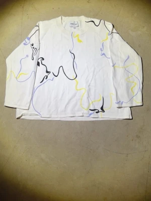 Bimba y Lola långarmad tröja - Vit långärmad t-shirt i bomull med abstrakta slingrande linjer i svart, gul och lila över hela plagget. Tröjan har en avslappnad passform och rund halsringning. Perfekt för dig som gillar unika och kreativa prints.