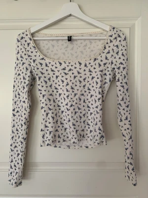 Vit långärmad figurnära blommig topp - Söt vit långärmad topp med små mörkblå blommor och ribbad struktur. Toppen har en fyrkantig/rund ringning med spetsdetalj längs kanten och är figurnära i modellen. Perfekt för dig som gillar romantiska och feminina plagg.