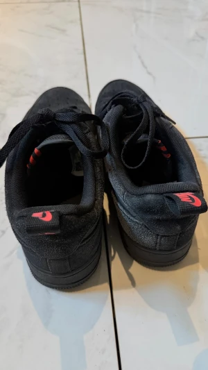 Svarta Nike sneakers med röd detalj - Snygga svarta sneakers från Nike i mocka med röda detaljer på hälen och tungan. Klassisk låg modell med svart sula och svarta skosnören. Perfekta för dig som gillar stilrena och sportiga skor.