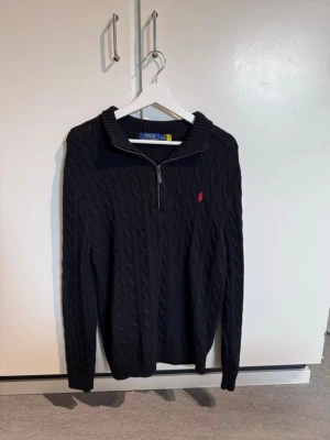 Svart kabelstickad tröja Polo Ralph Lauren - Kabelstickad svart tröja från Polo Ralph Lauren med halv dragkedja och hög krage. Tröjan har det klassiska röda logobroderiet på bröstet och ribbade muddar vid ärmslut och nederkant. Perfekt för en stilren och avslappnad look.