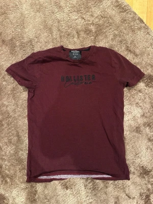 Vinröd Hollister t-shirt med tryck - Säljer en vinröd t-shirt från Hollister med svart broderad logga och diskret mönster. T-shirten har rund halsringning och korta ärmar. Perfekt för en avslappnad och stilren look.