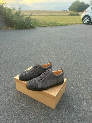 ÄKTA, helt nya Louboutins - Säljer ett par gråa sneakers från Christian Louboutin i mocka med svarta nitar på tån. Skorna har svarta snören, låg profil och en svart sula. Insidan är beige och designen är riktigt edgy och unik.