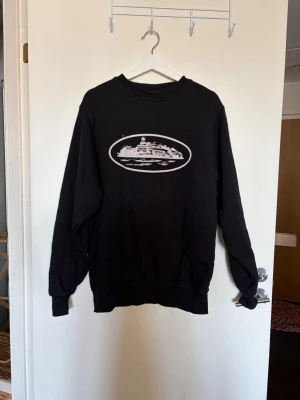 Svart sweatshirt Corteiz - Svart sweatshirt med rund hals och ett vitt tryck av en byggnad i oval ram på bröstet. Tröjan har långa ärmar och ribbade muddar vid ärmslut och nederkant. 