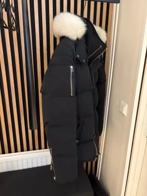 Svart dunjacka med pälshuva - Snygg svart dunjacka med stor fluffig vit pälskant på huvan och silvriga dragkedjor. Jackan har en patch på ärmen och flera praktiska fickor. Puffig modell som håller dig varm under kalla dagar. Perfekt för dig som vill ha både stil och funktion.