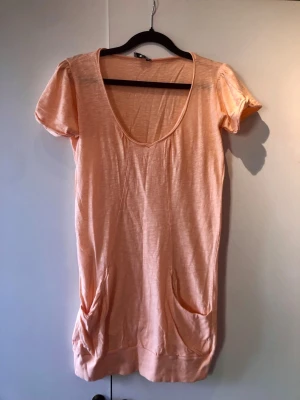 Bik Bok Lång Tröja / T-shirtklänning med Fickor Persiko – Stl S - Skön och lättburen lång tröja eller kort t-shirtklänning från Bik Bok (modell Frank) i en härlig persikoorange/rosa nyans. Tillverkad i mjuk och luftig bomull med djup urringning, söta rynkade ärmar, bred mudd i nederkanten samt två praktiska fickor framtill. Perfekt som ett avslappnat vardagsplagg eller över badkläderna på sommaren. Märke: Bik Bok Storlek: S Mått: Axelbredd ca 35 cm, längd från axeln ca 76 cm Material: 100% Bomull Skick: Mycket gott skick