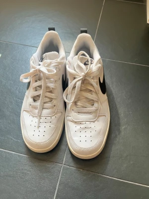 Vita Nike sneakers med svart swoosh - Snygga vita Nike sneakers med klassisk svart swoosh på sidan. Skorna har lågt skaft, vita skosnören och ventilerande hål på tån. Tillverkade i slitstarkt material med bekväm innersula och svart detalj på hälen. Perfekta för en clean och sportig look.