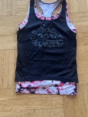 Svart och blommigt träningslinne SOC - Säljer ett snyggt träningslinne från SOC i storlek 134/140. Linnet är svart framtill med texten 'Soc Sports Wear' och har blommigt mönster i rosa, vitt och svart på kanter och baksida. Perfekt för gym, löpning eller annan träning.