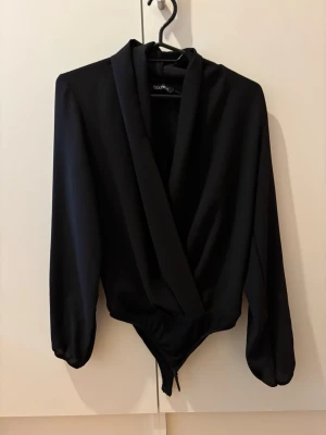 Svart långärmad body med omlott - Stilren svart body med långa ärmar och djup omlott-ringning.  ger en elegant känsla. Perfekt att matcha med både jeans och kjol för en trendig look.
