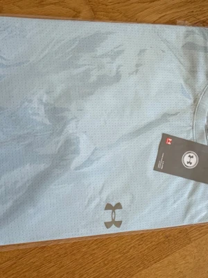 Ljusblå träningströja Under Armour - Snygg ljusblå träningströja från Under Armour i ventilerande meshmaterial. Perfekt för gymmet eller löprundan med sin lätta och snabbtorkande kvalitet. Rund halsringning och diskret logga på bröstet ger en clean sportig look.
