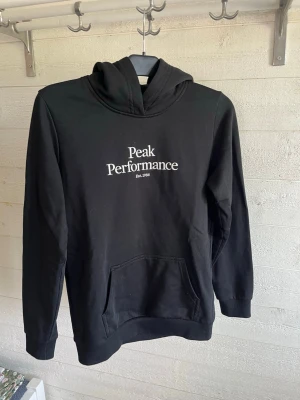 Svart hoodie från Peak Performance - Snygg svart hoodie från Peak Performance med vit broderad logga på bröstet. Klassisk känguruficka framtill och huva. Tillverkad i mjukt material som känns skönt mot huden. Perfekt för en avslappnad och sportig stil.