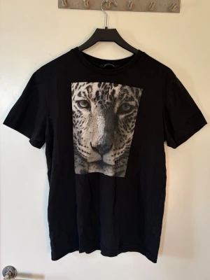 Tröja med tryck - Snygg svart t-shirt från The Cool Elephant med ett stort leopardtryck i svartvitt på bröstet. Klassisk rund hals och korta ärmar. Perfekt för dig som gillar djurmotiv och vill sticka ut lite extra.