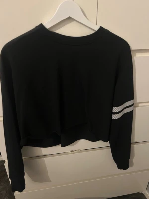 Svart cropped hoodie från Stronger - Säljer en svart cropped hoodie från Stronger med vita ränder på ena ärmen och loggan 'STRONGER' tryckt framtill. Tröjan har långa ärmar och en avslappnad passform. Perfekt till träning eller chill dagar.