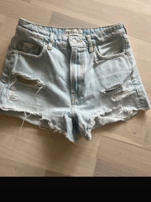 Ljusa denimshorts från Zara - Säljer ett par ljusblå denimshorts från Zara med hög midja och slitna detaljer. Shortsen har fransig kant, klassiska fem fickor och dragkedja med knapp. Perfekta för sommaren och ger en avslappnad, trendig look.