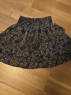 Blommig kortkjol från H&M - Supersöt kortkjol från H&M med blommigt mönster i svart, gul, lila och blå. Kjolen har en bred resår i midjan som ger en bekväm passform och ett flowy fall. Perfekt för dig som gillar färg och mönster! Har också lite trådar som sticker ut från sömmarna pga andvändning.