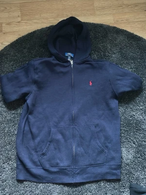 Mörkblå hoodie från Polo Ralph Lauren - Mörkblå hoodie från Polo Ralph Lauren med dragkedja framtill och känguruficka. Klassisk röd logga broderad på bröstet. Tillverkad i mjukt bomullsmaterial som känns skönt mot huden. Perfekt för en avslappnad och stilren look. XL på BarnStorlek, XS herrstorlek