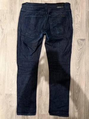 Nudie Jeans Grim Tim Dry Mörkblå W34/L32 - Slim Straight Fit - Nudie Jeans Grim Tim ”Dry Navy Dips” mörkblå Dry-jeans med slim straight fit-passform, normal midja och raka ben, storlek W34/L32, midja 43 cm, ytterbenslängd 99,5 cm, benöppning 17 cm, modellen är 185cm & väger 65kg, i fint skick.