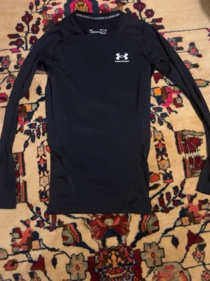 Under Armuor - Säljer en svart långärmad kompressionströja från Under Armour med HeatGear-teknologi. Tröjan har en tight passform och logga på bröstet. Perfekt för träning eller löpning när du vill ha något som andas och sitter skönt.