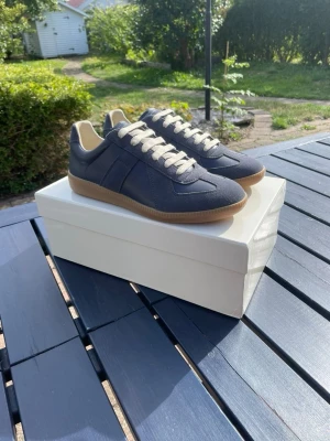 Maison Margiela - Snygga mörkblå sneakers med beige detaljer och klassisk gummisula. Skorna har vita snören och en stilren design med mockaliknande material på ovandelen. Perfekta för dig som gillar en clean och tidlös look.
