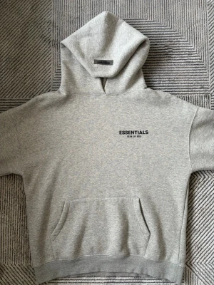 Grå Essentials Fear of God hoodie - Snygg grå hoodie från Essentials Fear of God med svart logga på bröstet och stor text på ryggen. Klassisk känguruficka framtill och stor huva. Tillverkad i mjukt bomullsmaterial, perfekt för en avslappnad streetwear-look.