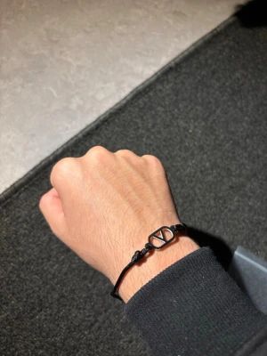Valentino armband - Snyggt svart armband med justerbart band och en stilren metallberlock i form av en rektangel med ett geometriskt mönster. Perfekt accessoar för dig som gillar minimalistisk och modern stil. Passar både till vardagslooken och när du vill addera något extra.