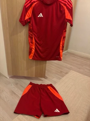 Rött Adidas träningsset med orange detaljer - Säljer ett sportigt träningsset från Adidas i rött med orange detaljer. Setet består av en t-shirt och shorts, båda med vita Adidas-loggor. Materialet är lätt och andas, perfekt för träning eller match. Snygg design med kontrastfärgade ränder på axlar och sidor.