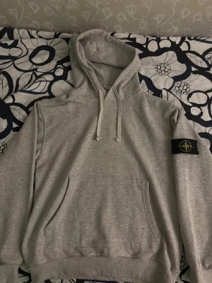 Grå hoodie med patch på ärmen. Storlek M men passar även som S. - Snygg grå hoodie med huva och dragsnören samt en stor magficka framtill. På vänster ärm sitter en svart patch med gult och grönt broderat märke. Hoodien har ribbade muddar vid ärmslut och nederkant. Perfekt för en avslappnad och trendig look.