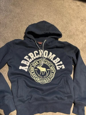 Mörkblå hoodie från Abercrombie - Mörkblå hoodie med stor vit Abercrombie-logga och tryck framtill. Klassisk huva med dragsko och känguruficka. Mjuk bomullsblandning och ribbade muddar vid ärmslut och nederkant. Perfekt för en avslappnad och sportig stil. PRUTBART!