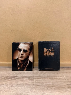 The Godfather DVD-box samlingsutgåva - Säljer en The Godfather DVD Collection box med svart och guld design. Boxen innehåller flera DVD-skivor med filmerna och tillhörande material. Omslaget har porträtt och klassisk logotyp. Perfekt för dig som gillar maffiafilmer och vill ha hela trilogin samlad i ett snyggt fodral.