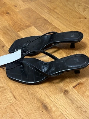 Svarta flip flop kitten heels  - Stilrena svarta sandaletter från H&M med låg klack och fyrkantig tå. Remmarna är tunna och ovandelen har ett croco-mönstrat konstläder som ger en trendig look. Perfekta för dig som gillar minimalistisk och modern stil. Passar storlek 36-37. Prislapp kvar.