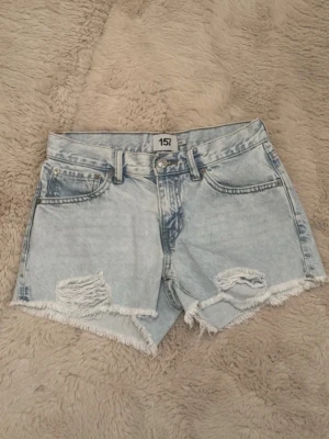Ljusblå denimshorts med slitningar - Säljer ett par ljusblå denimshorts med råa kanter och slitna detaljer framtill. Shortsen har fem fickor, bälteshällor och knappgylf. Perfekta för sommaren och ger en avslappnad vibe till din outfit.