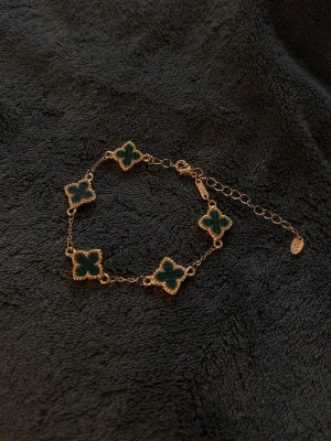 Guldarmband med gröna fyrklöver - Snyggt armband i guldton med fem gröna fyrklöverformade hängen. Kedjan är tunn och har justerbar längd med ett litet runt märke vid låset. Perfekt för dig som gillar unika detaljer och stilrena accessoarer.